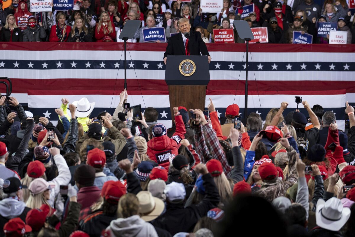 President Trump addresses the crowd from a stage decorated in red, white and blue while a crowd stands behind him.