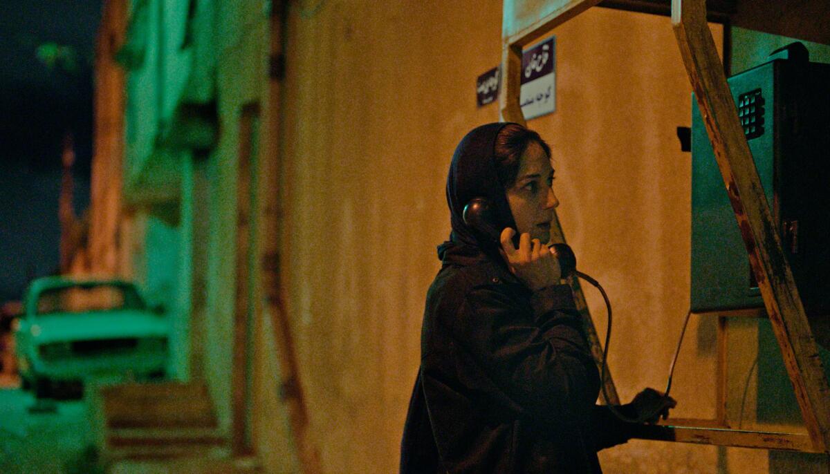 A woman stands outside at night on an empty city street talking on a public phone.