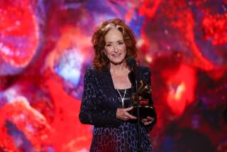 A woman with curly red hair wearing a suit and holding a trophy in front of a microphone on a stage