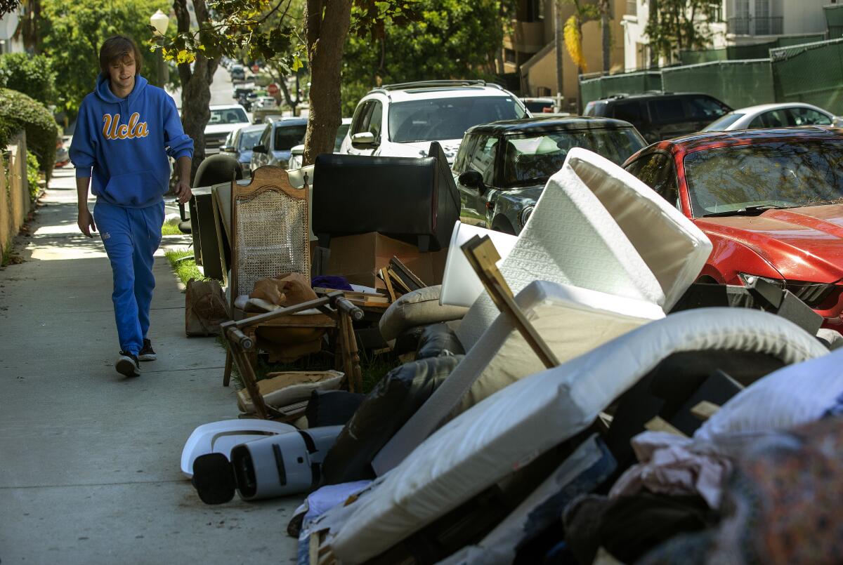 A young man wearing a UCLA walks past discarded furniture on the sidewalk.
