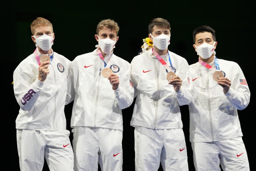 Four young men in face masks and USA warmup suits pose while holding up their medals.