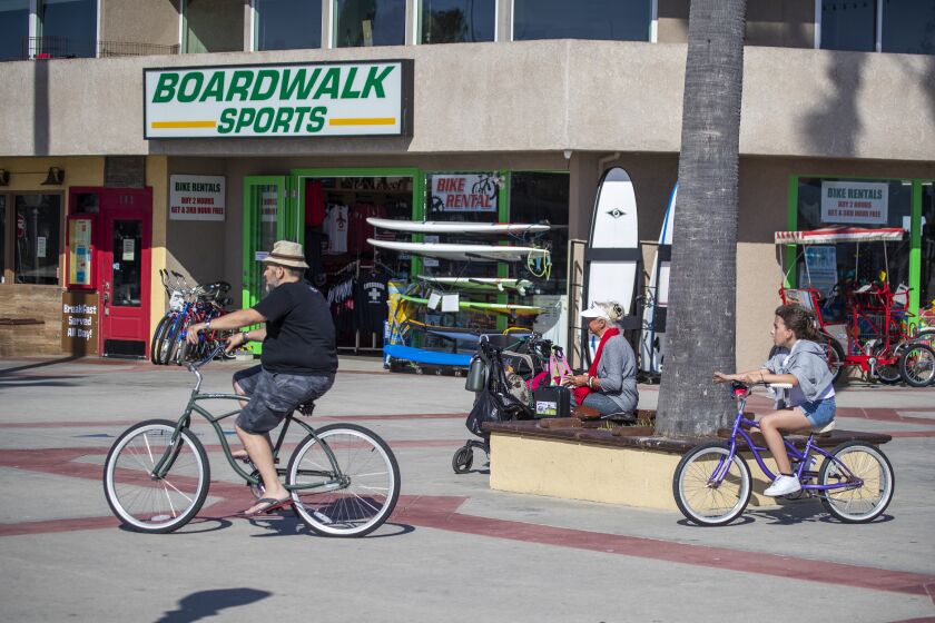 NEWPORT BEACH, CA -- MONDAY, MAY 4, 2020: A bicyclist and pedestrians pass the closed Boardwalk Sports store on the boardwalk in Newport Beach, CA, on May 4, 2020. The business is only renting bicycles but is hoping to fully open after Gov. Newsom's announcement Monday that some businesses will be able to reopen. (Allen J. Schaben / Los Angeles Times)