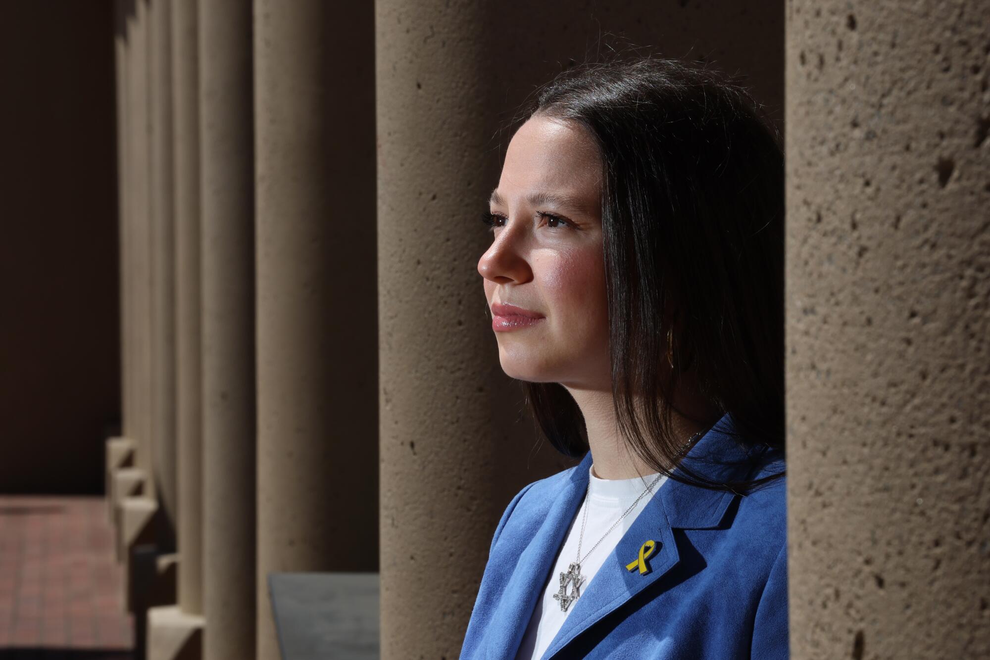 Alyssa Wallack stands between building columns while wearing a Star of David necklace and yellow ribbon lapel pin