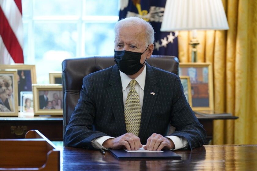 President Biden sitting behind his desk in the Oval Office, wearing a protective mask.