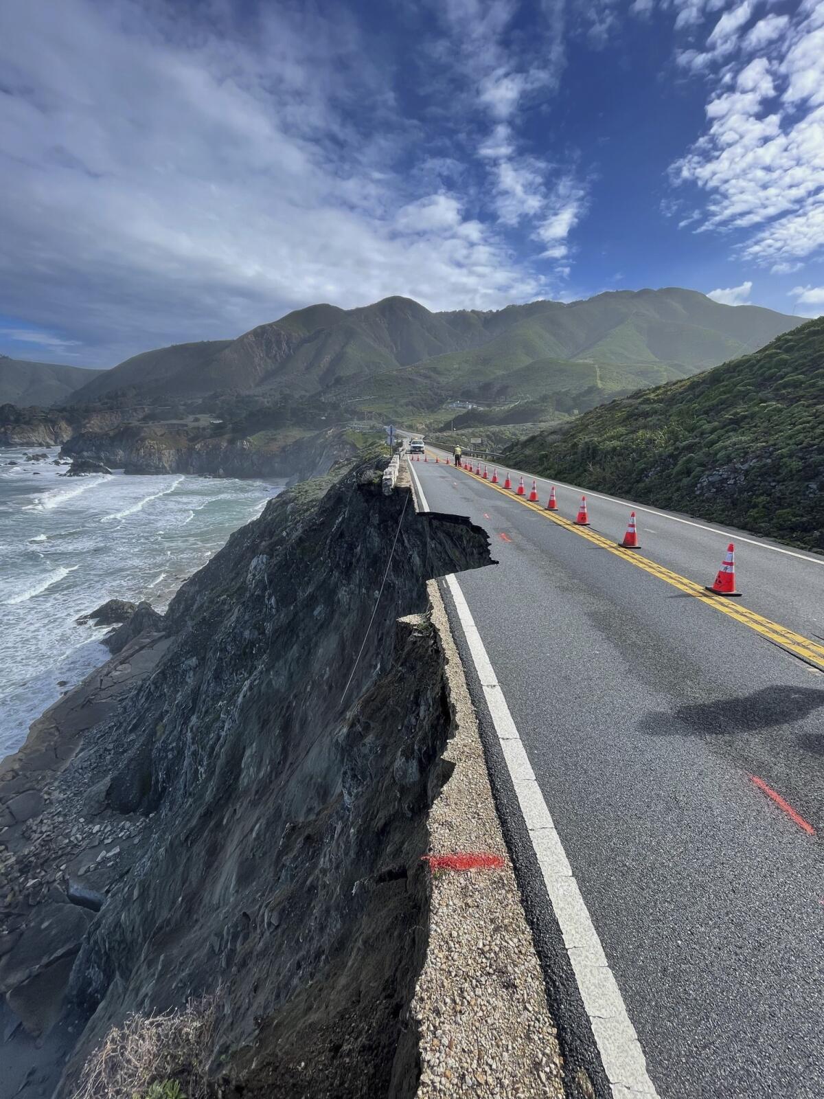 A coastal roadway has a chunk of asphalt missing. In the background is ocean, mountains and a blue sky with clouds.
