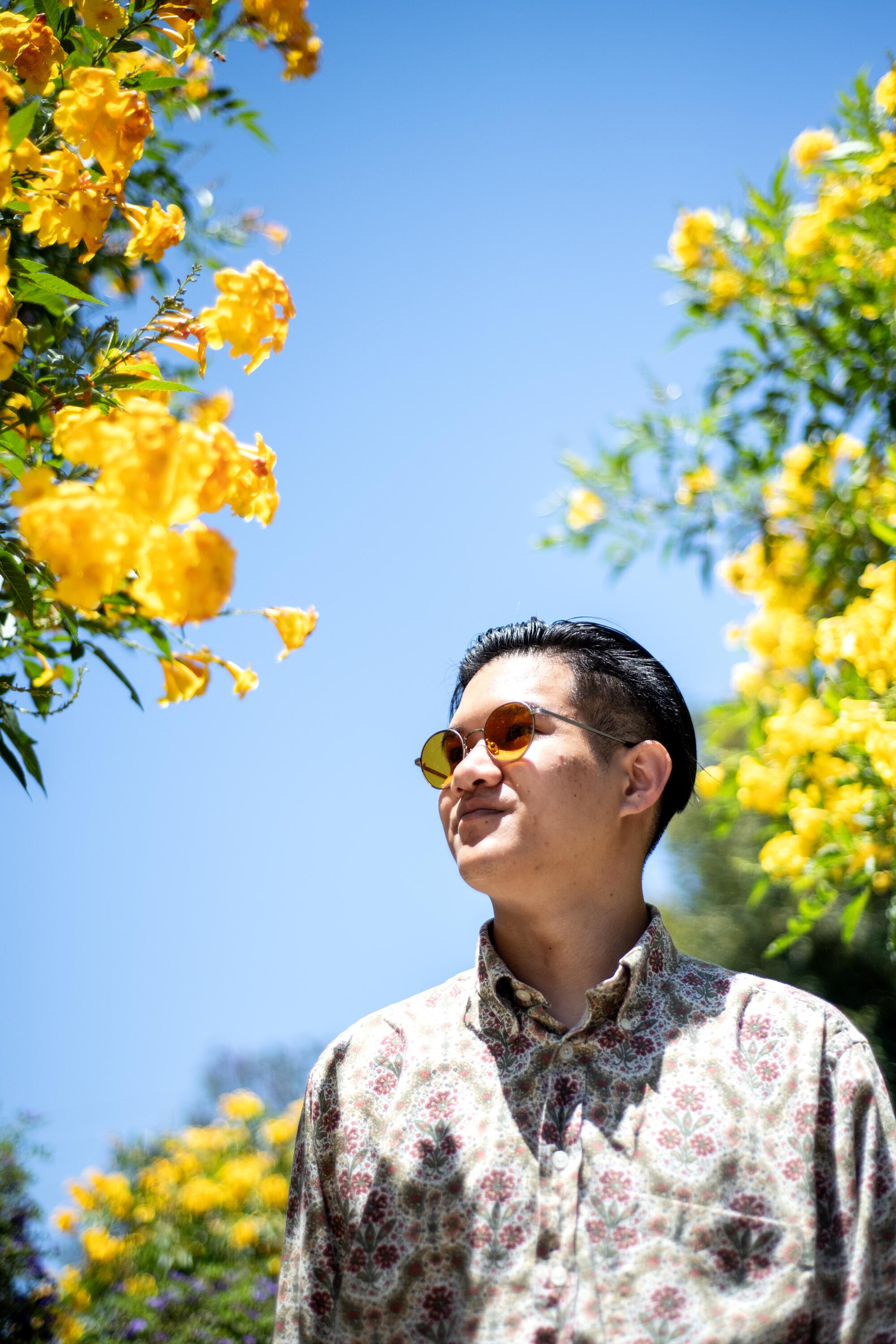 A man in a boldly patterned shirt and sunglasses stands amid yellow blooming trees.