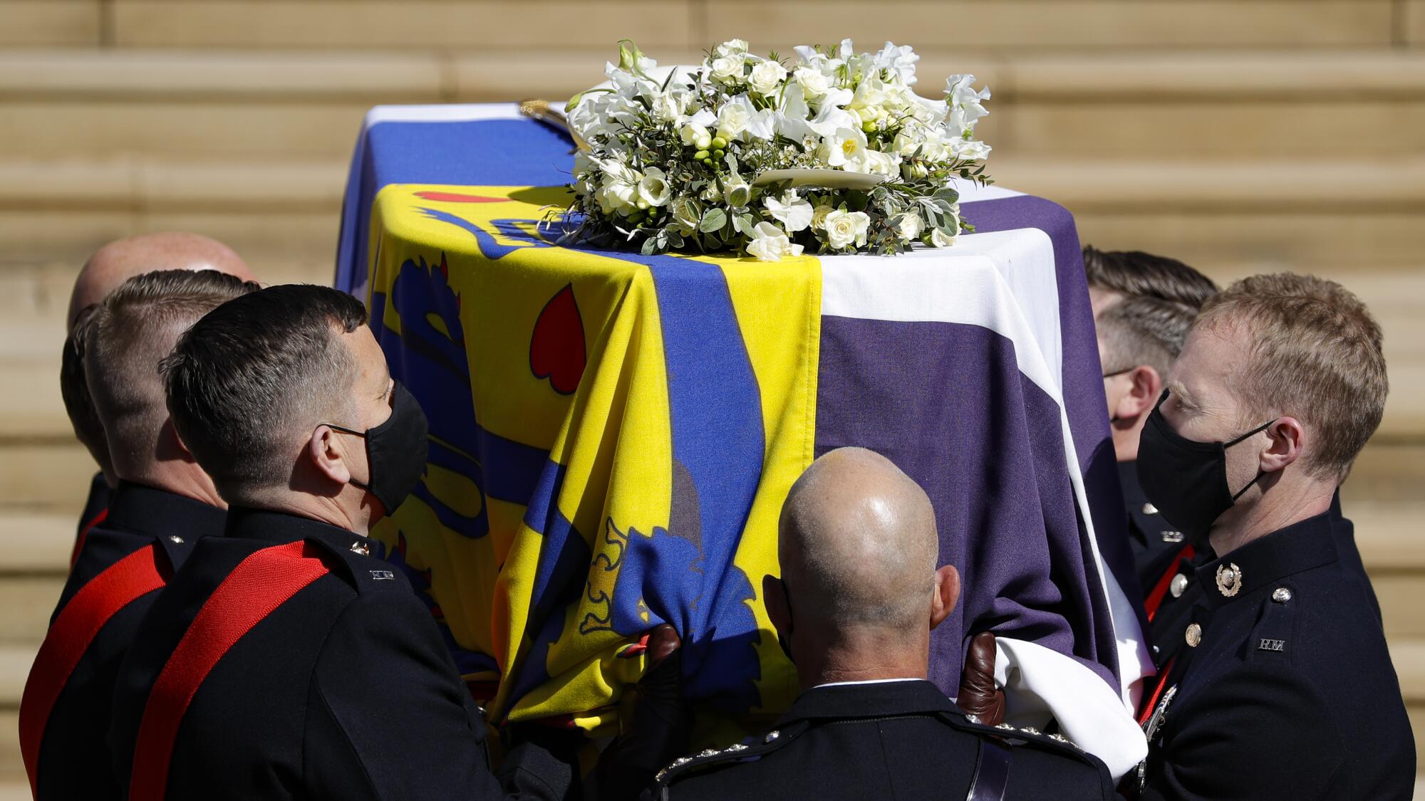 A closeup of the coffin with a multicolored cloth covering and flowers on it as pallbearers carry it up stairs.