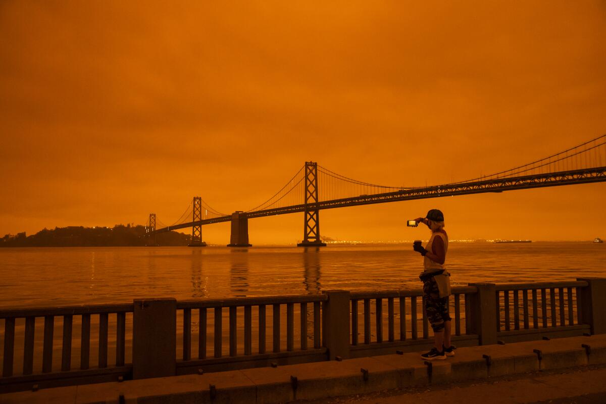A person photographs the San Francisco Bay Bridge under orange skies