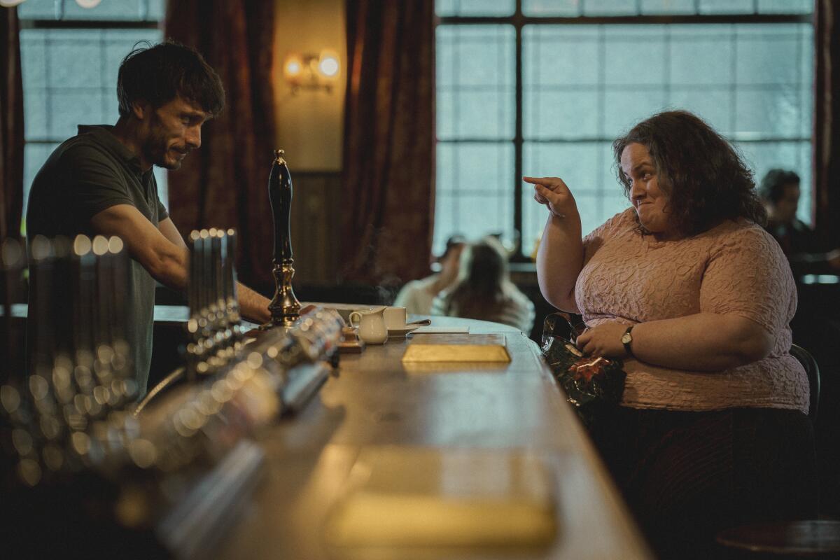 A woman sits at a bar pointing at the bartender
