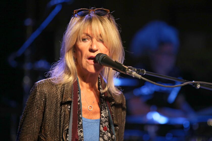 Christine McVie sings into a mic while wearing a brown jacket and blue shirt with sun glasses atop her head