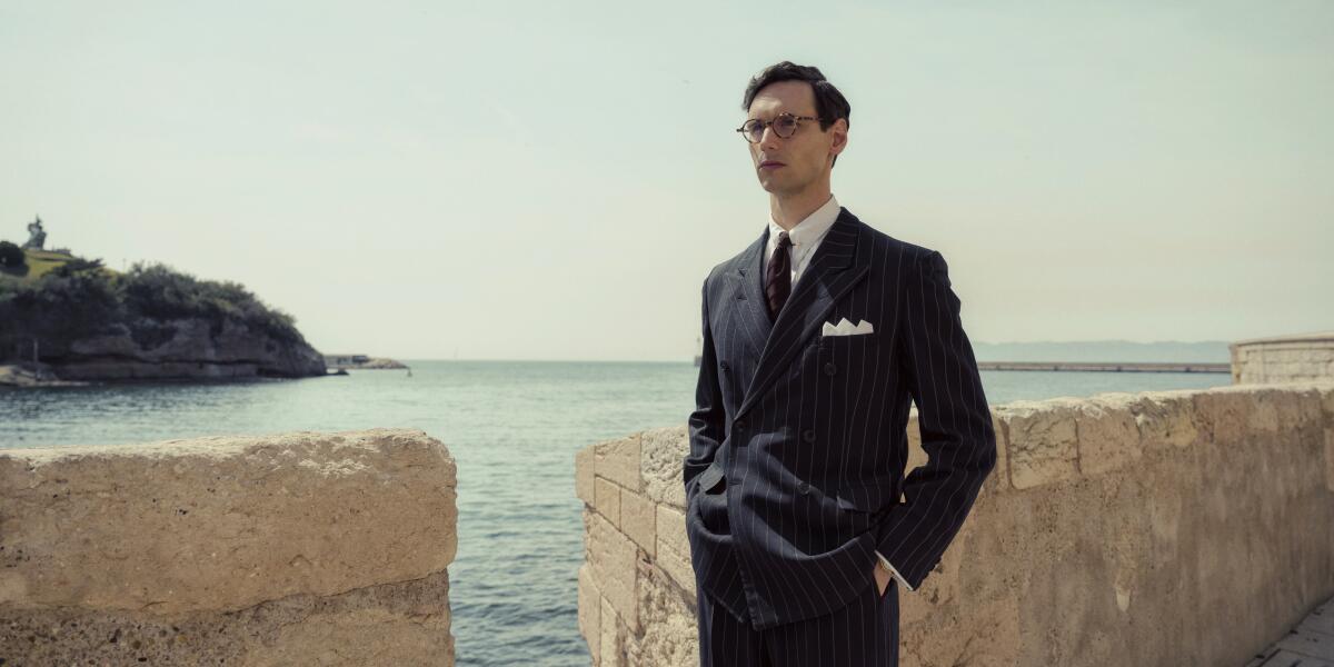 A man in a 1940s-style suit stands near a sea wall in a scene from "Transatlantic."