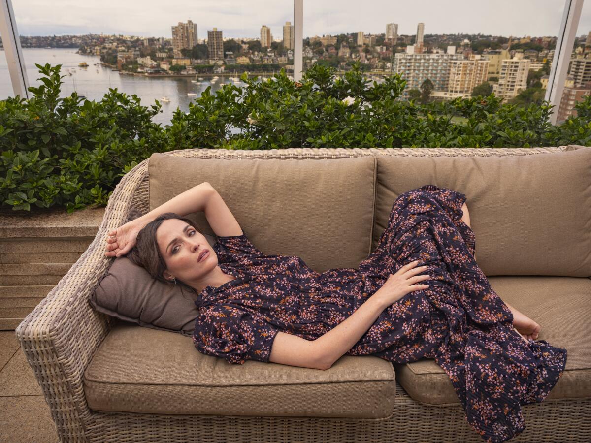 Rose Byrne lies on a rattan sofa. In the background, through windows, is a city skyline.
