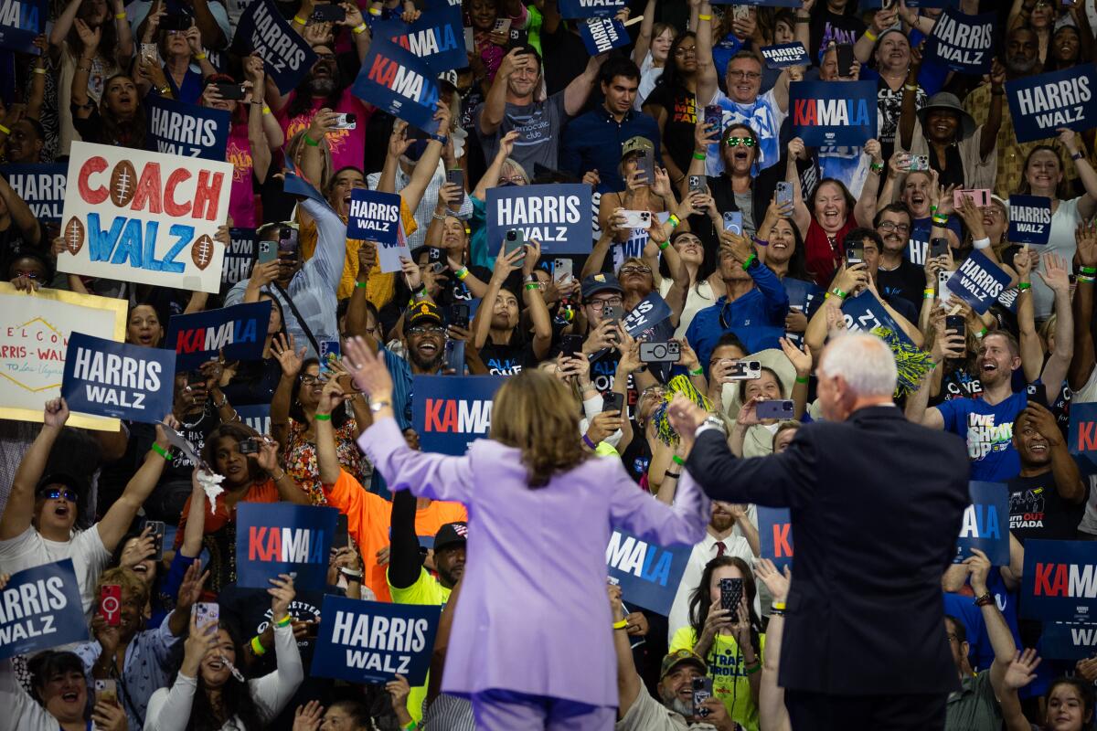 Kamala Harris and Tim Walz, from behind, wave at a crowd raising campaign signs, one reading "Coach Walz" with footballs