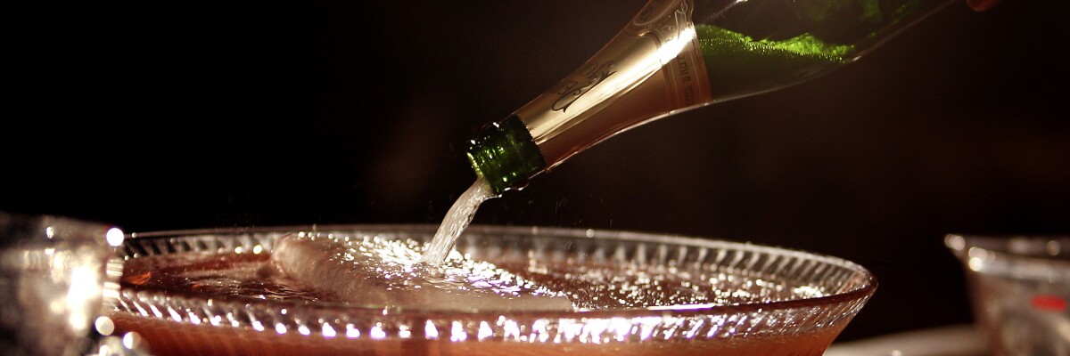 Champagne is poured from a bottle into a bowl full of reddish-purplish liquid with a large block of ice floating in it. 