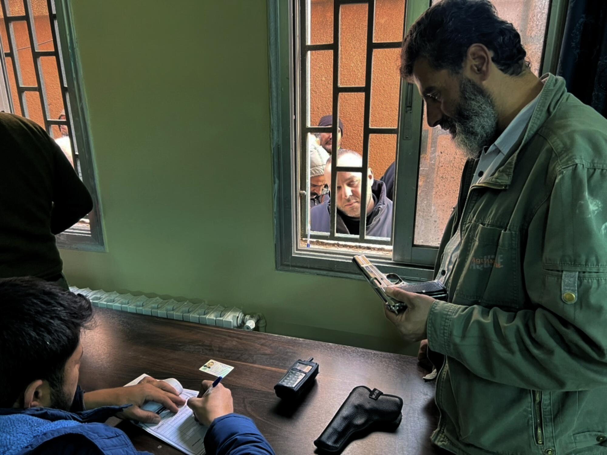 A man seated at a desk jots down information in a book as a man stands next to him holding a gun