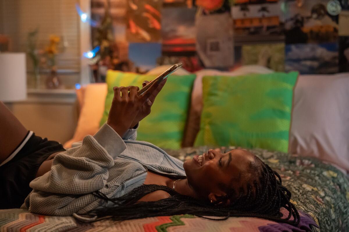 A teenage girl lays on her back on her bed reading on her phone.