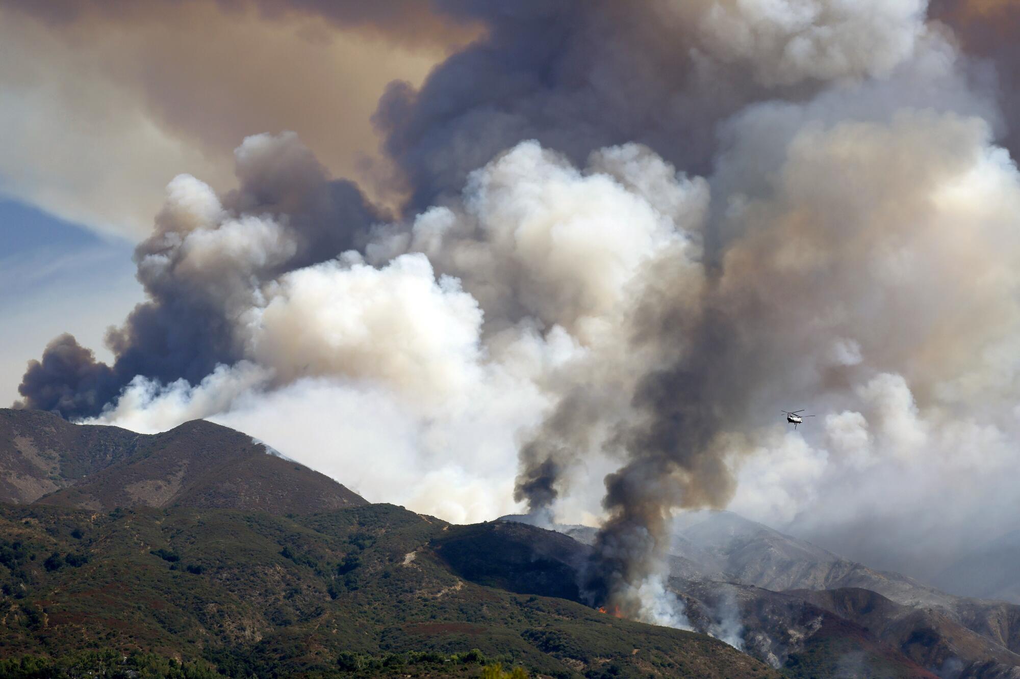 A firefighting helicopter is dwarfed by a giant column of gray smoke rising from a mountain.