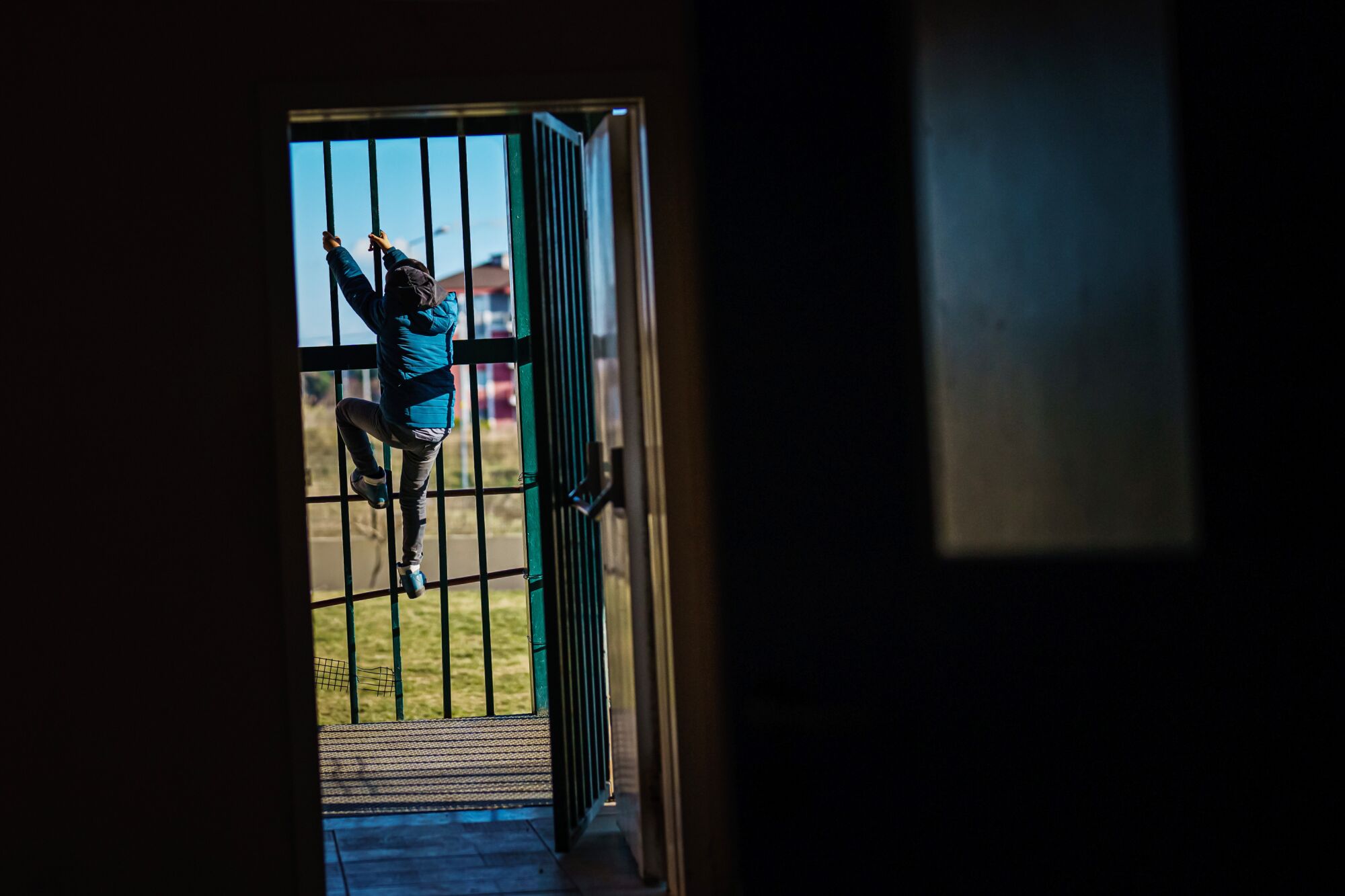 Seen through an open door, a child climbs a tall metal fence.