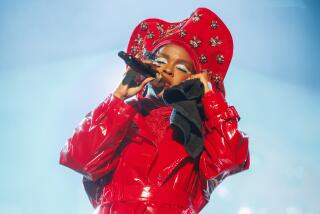 Lauryn Hill performing while holding a mic to her mouth and closing her eyes, dressed in a red jacket