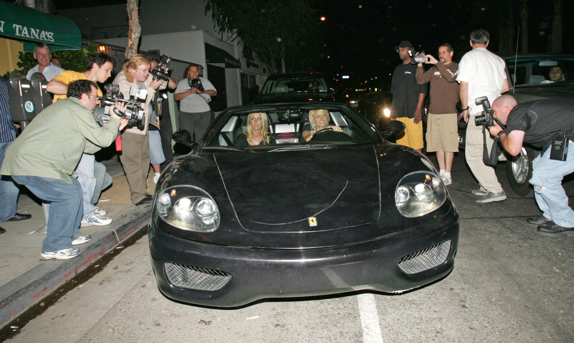 Nicky and Paris Hilton driving a black sports car with lines of photographers on either side of them