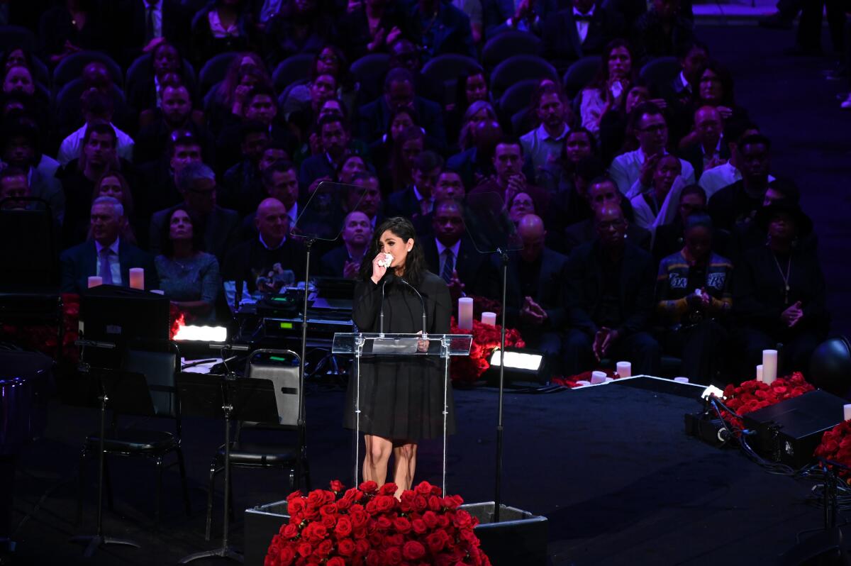 Vanessa Bryant holds a tissue to her face while speaking on a stage decorated with roses