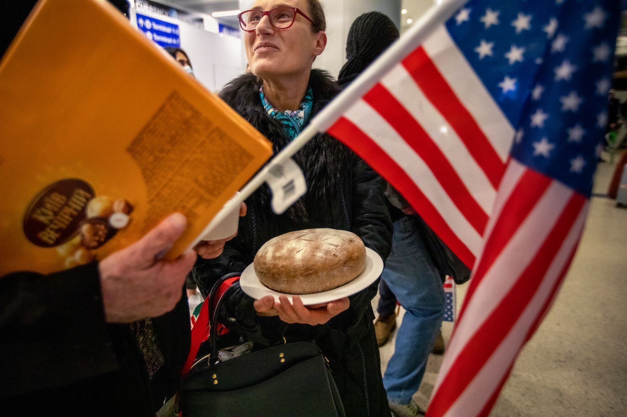 A woman stands, framed behind an American flag, holding a plate with bread on it.