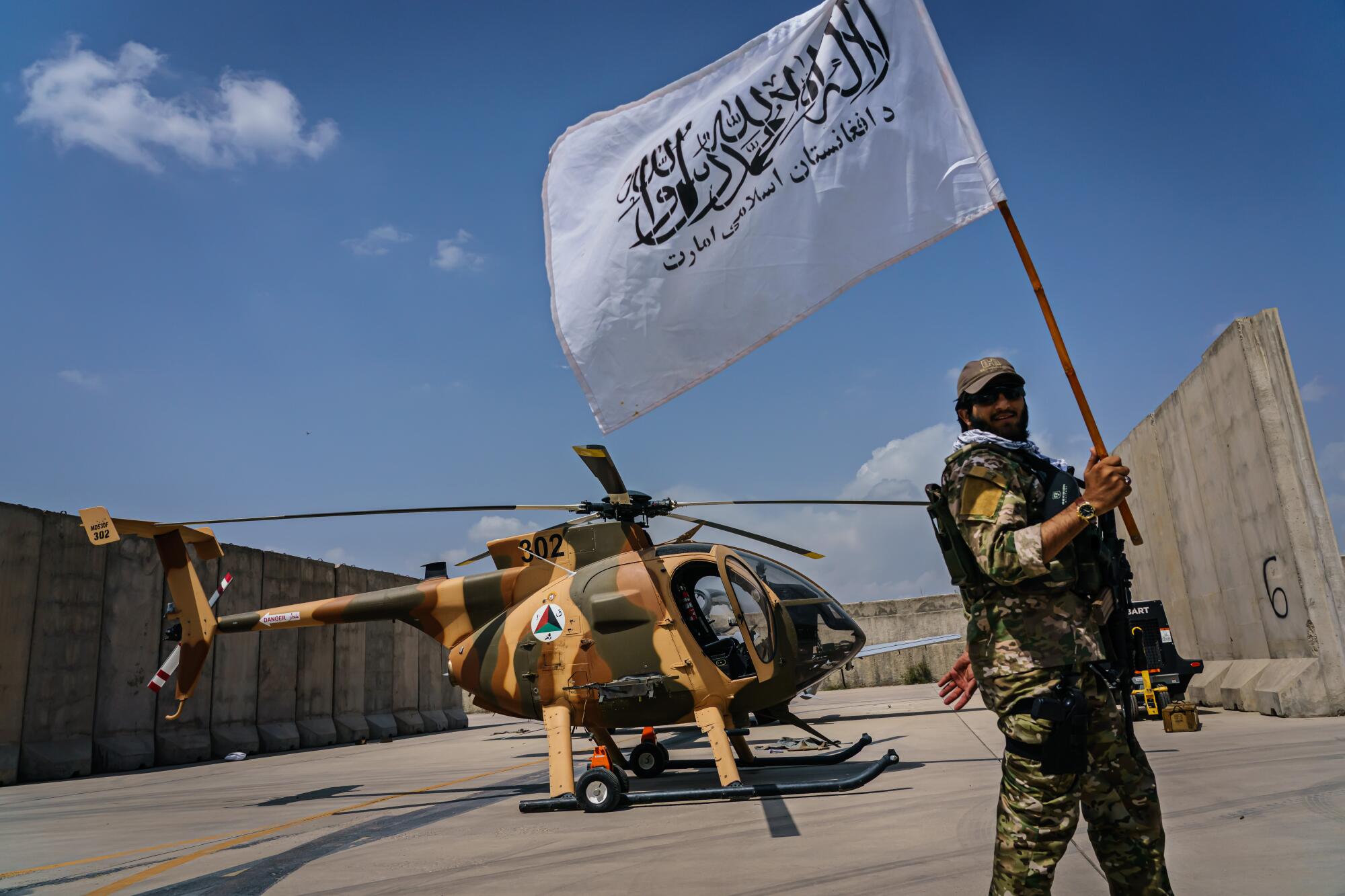 A Taliban fighter with white Taliban flag in front of a camouflage helicopter 