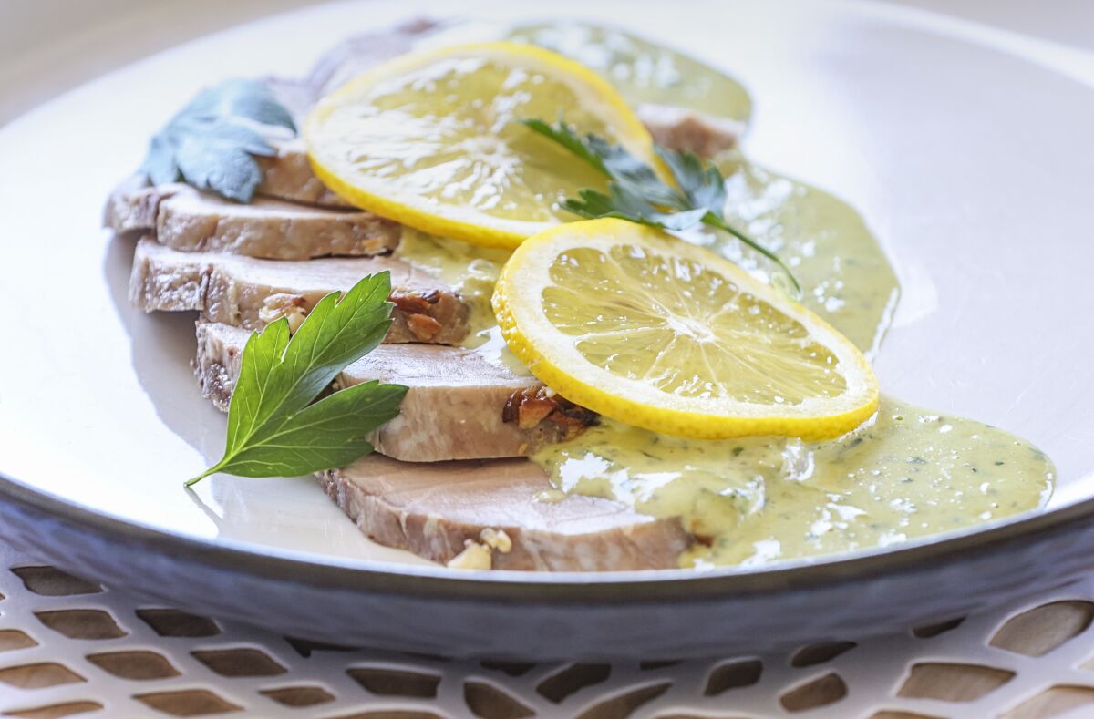 Slices of Pork Tenderloin Tonnato on a plate are topped with a tuna sauce and slices of lemon.