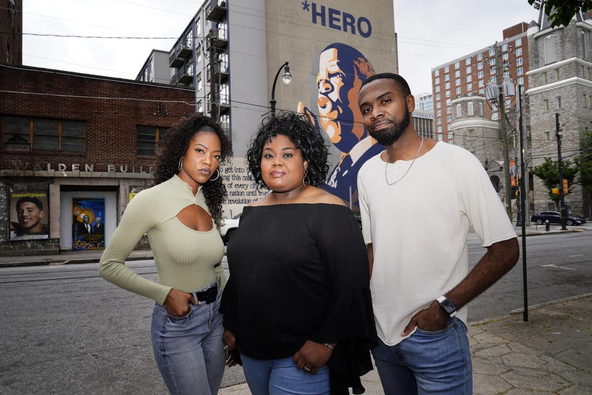 Three people pose on a sidewalk, a mural behind them 