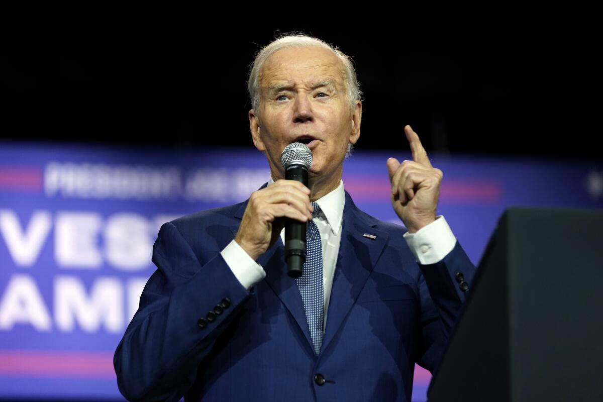 President Biden is seen holding a microphone and gesturing with his left hand as he speaks.