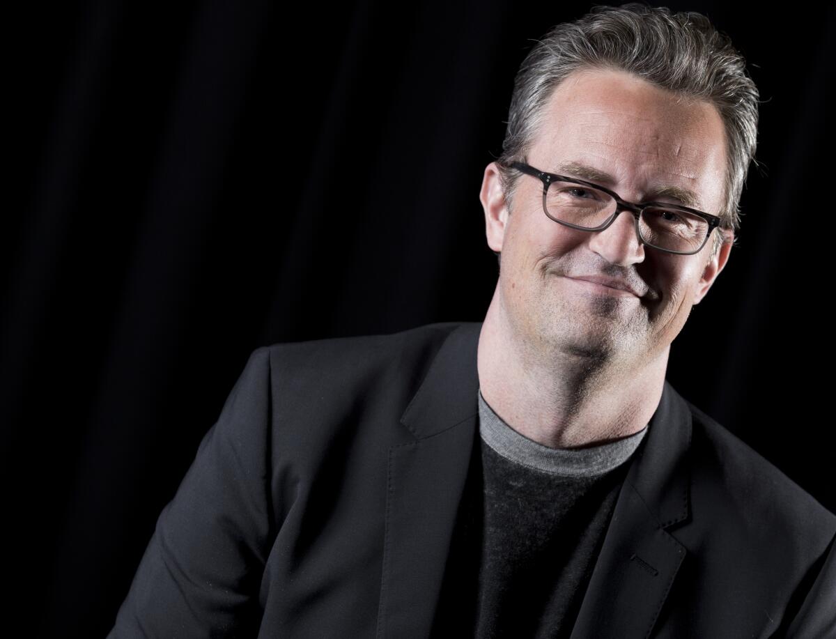 Matthew Perry in glasses, a shirt and black suit jacket.