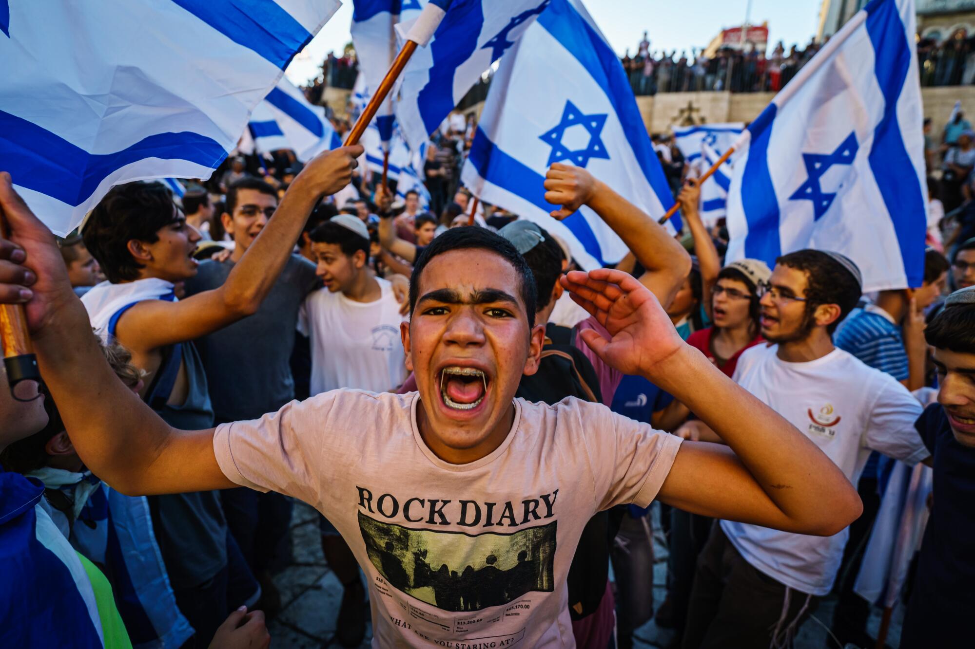 A young man with braces and wearing a "Rock Diary" T-shirt yells amid other young men waving Israeli flags.