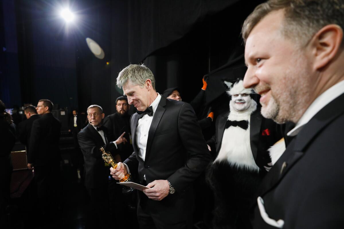 Dominic Tuohy and Greg Butler, winners of the visual effects Oscar for “1917” walks with James Corden as a cat backstage at the 92nd Academy Awards.