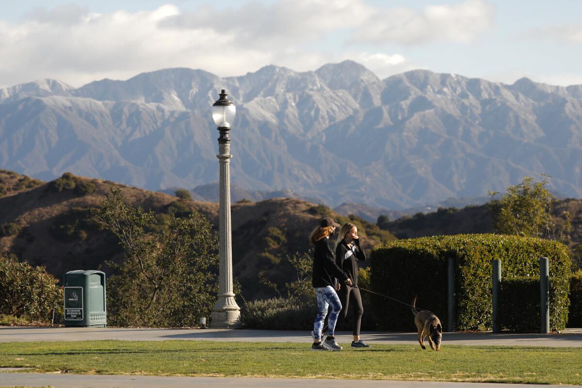 Two women and a dog walk with mountains in the background.