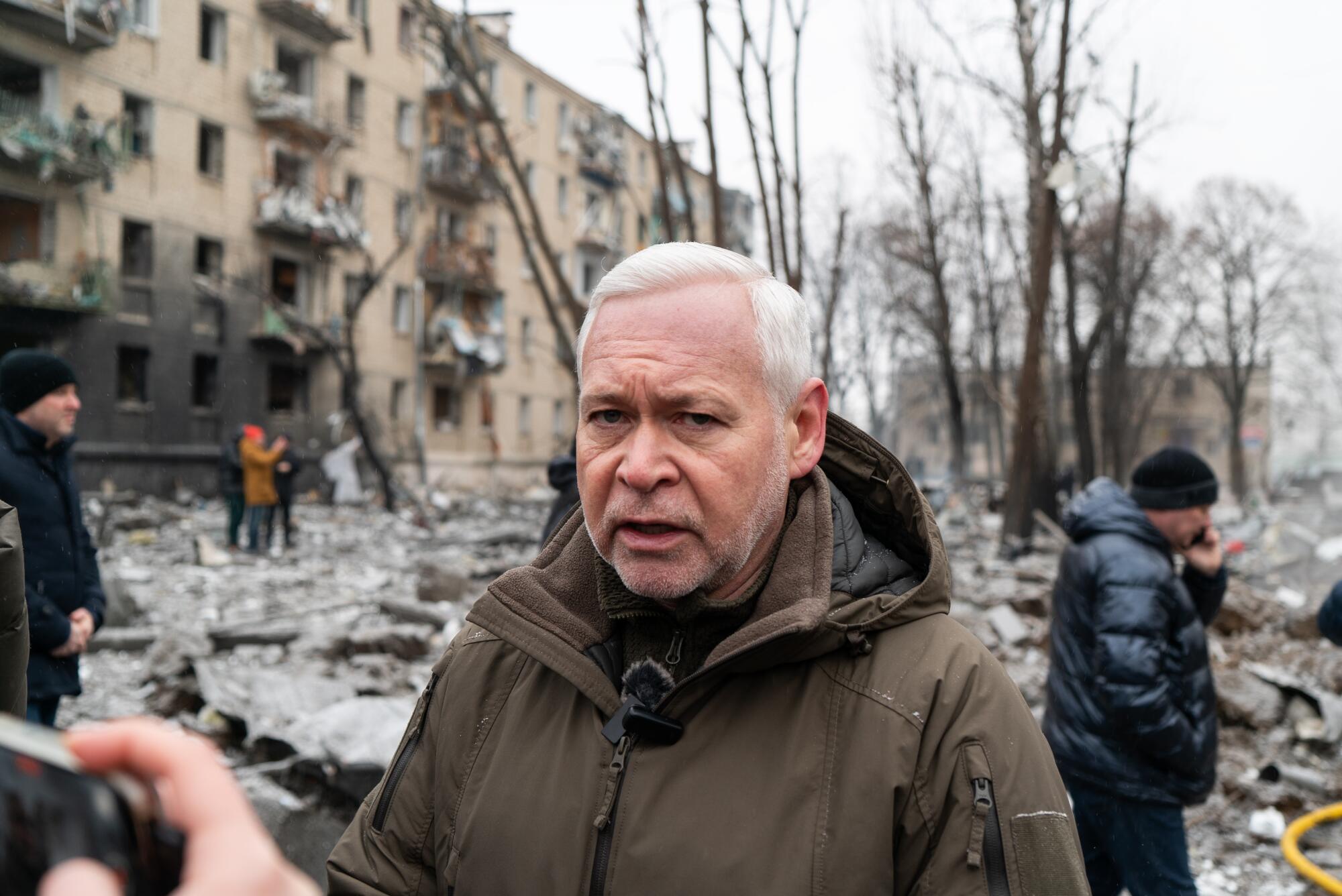 一名头发花白的男子，身穿棕色连帽夹克，背景是一栋受损的建筑，上面有烧焦的痕迹。