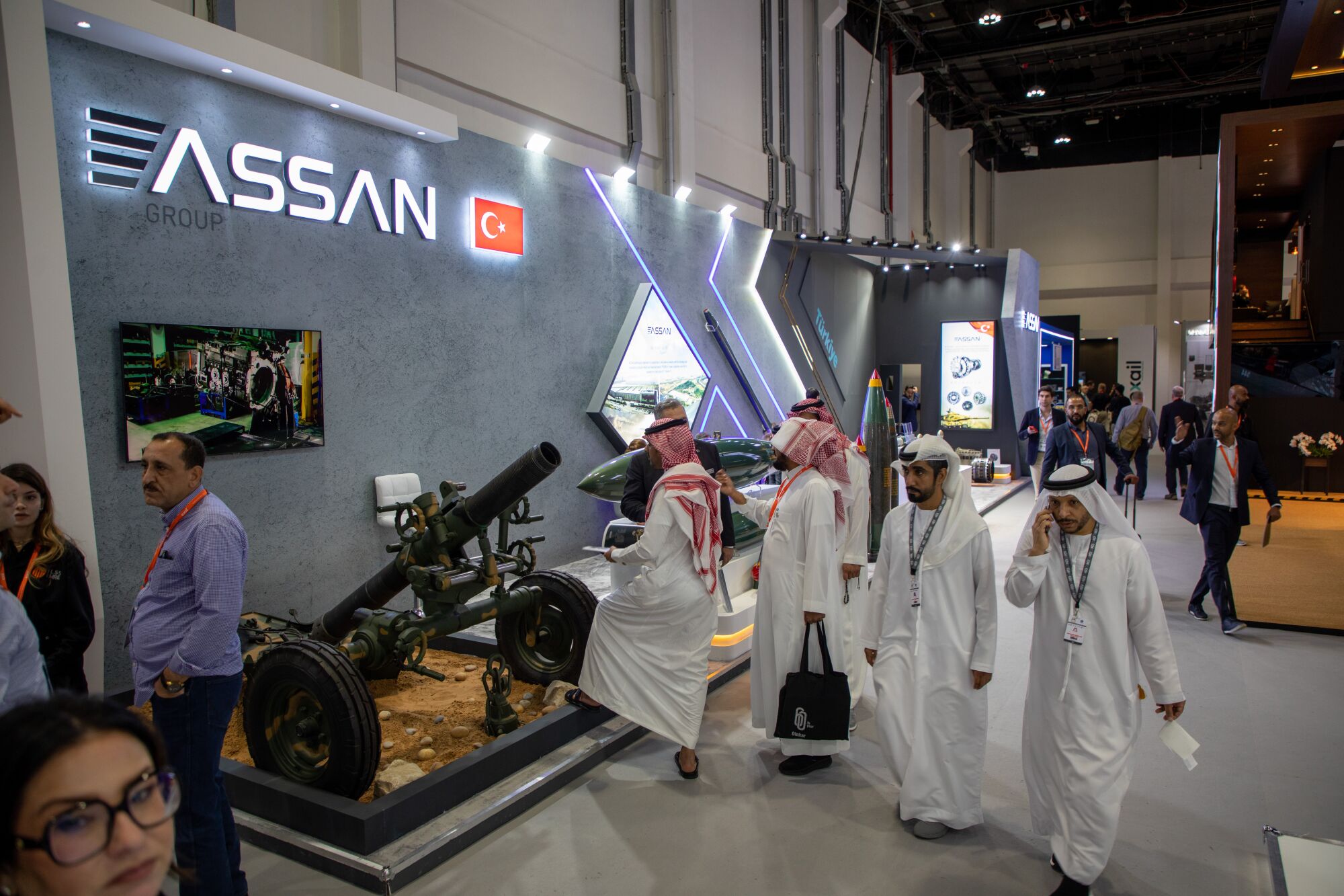 Several men in white robes and headdresses walk near a display featuring a weapons launcher