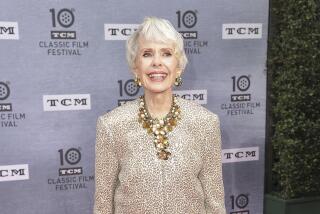 A woman with short white hair in a gold jacket and pants, wearing a large necklace and posing with her hands at her sides