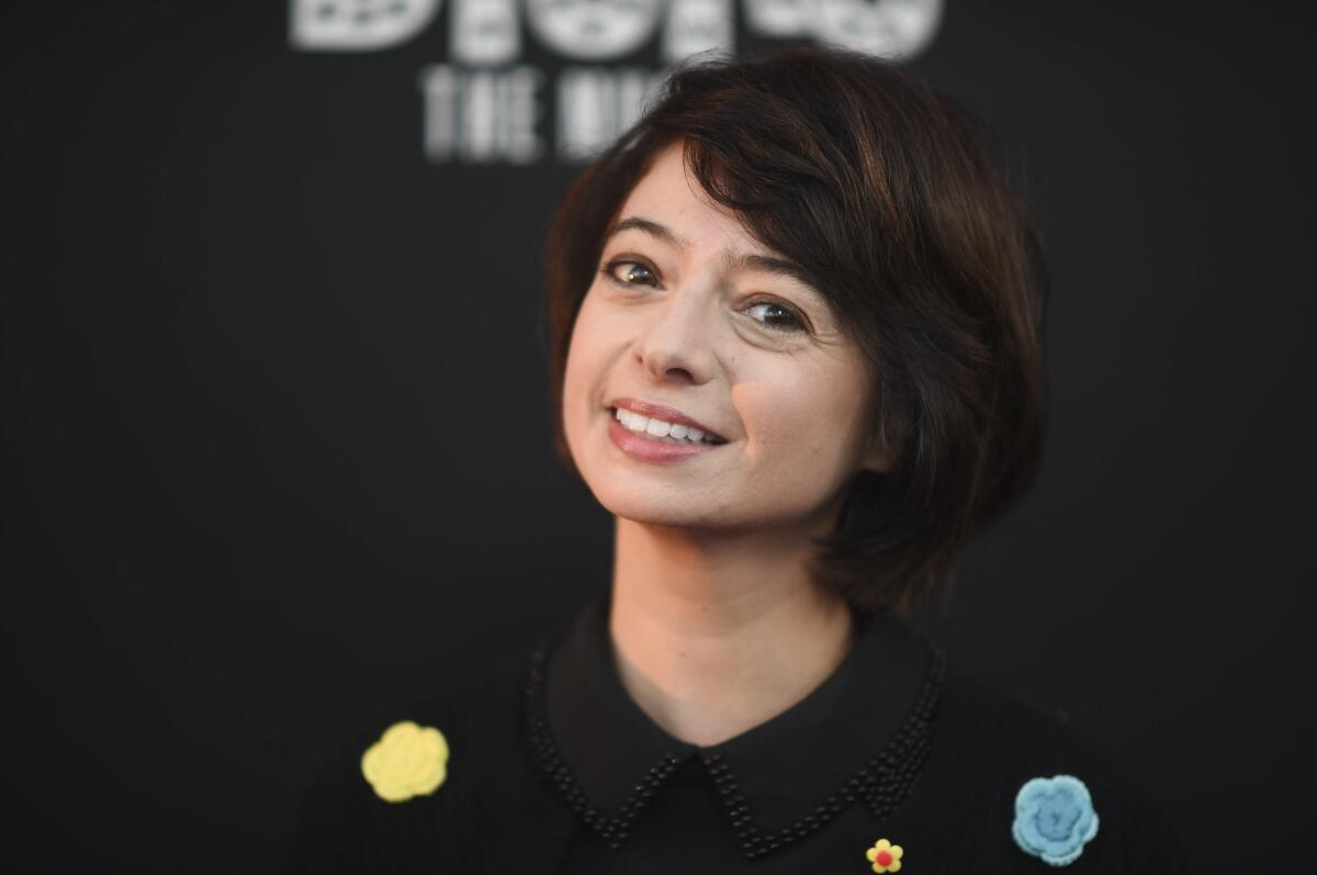 Kate Micucci wears a black sweater with blue and yellow knit flowers on it and poses for photos