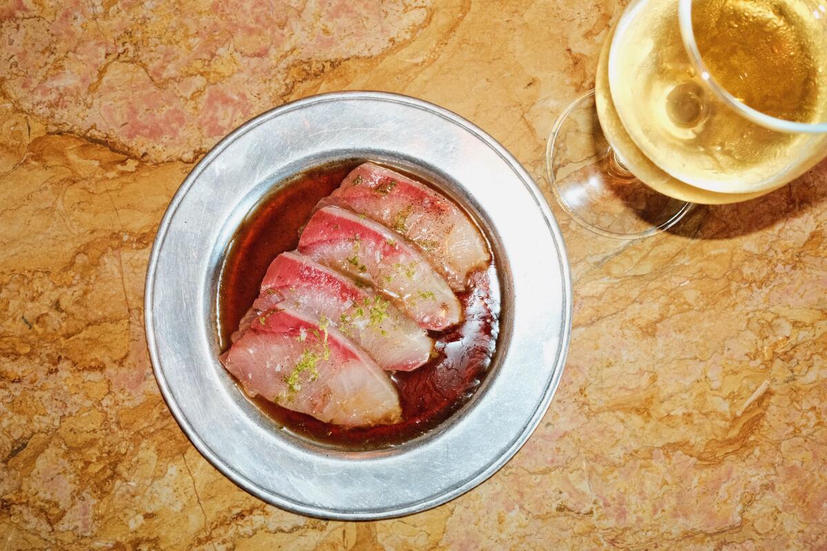 A dish of hamachi crudo on a silver plate next to a glass of white wine