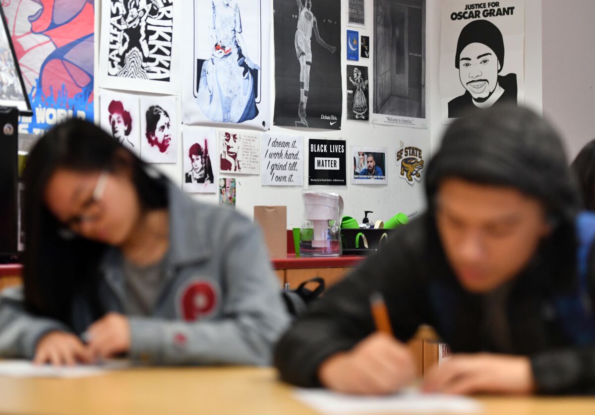 A female and male high school student, out of focus, work at desks in front of posters featuring Black Lives Matter, Oscar Grant, a Golden State Warrior and more.