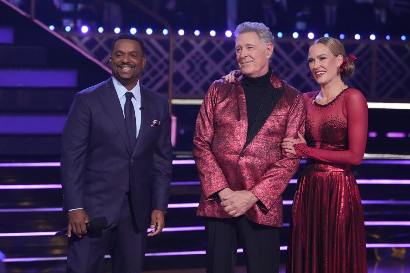 Alfonso Ribeiro in a blue suit, Barry Williams in a red suit and Peta Murgatroyd in a red dress, all standing on a stage