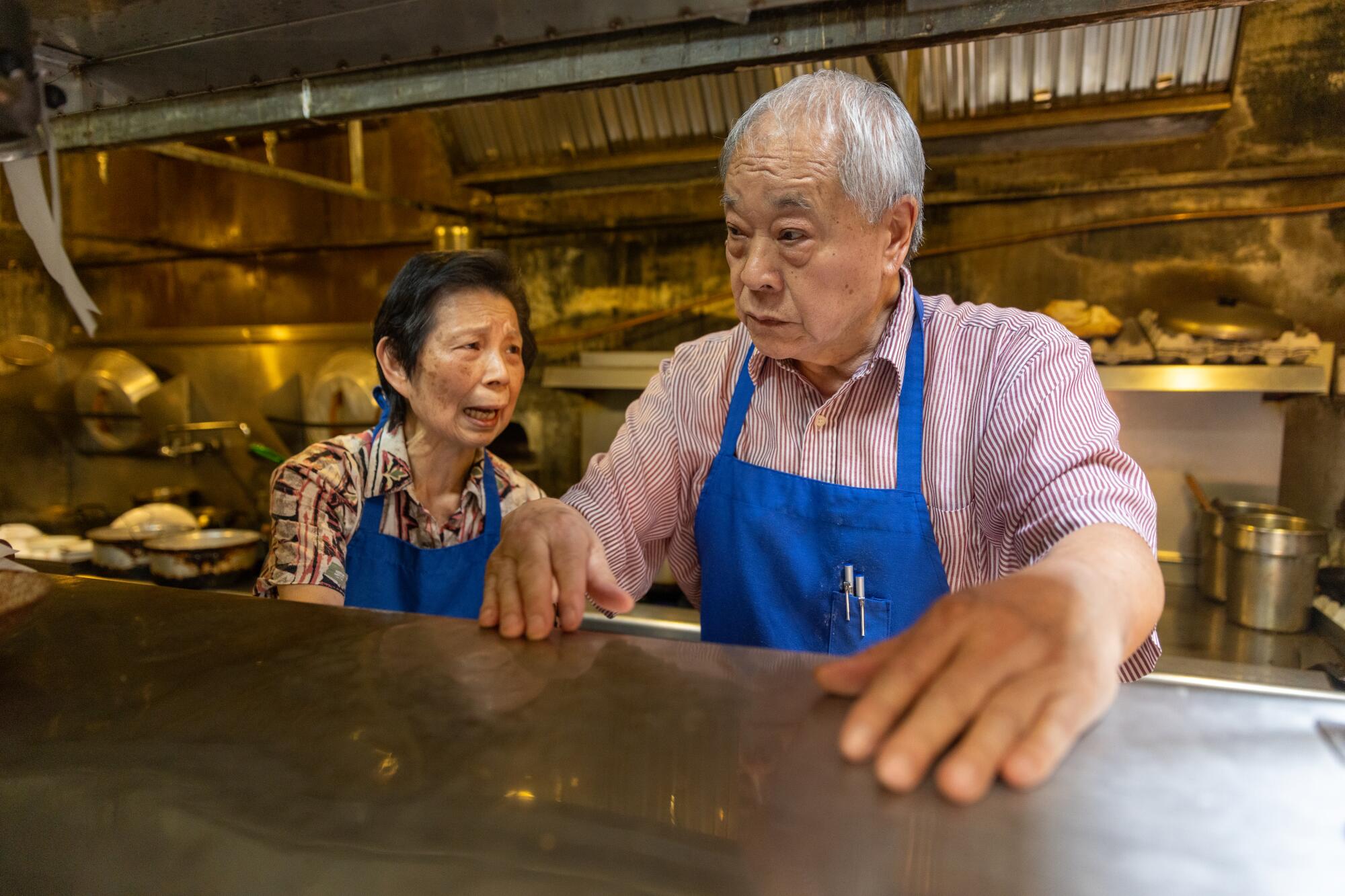 A husband and wife wearing blue aprons prepare meals in a restaurant kitchen. 
