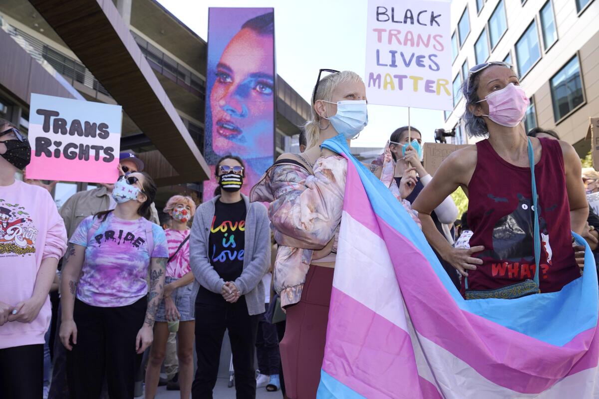 People stand outside wearing pink and light blue and holding signs that read "transgender rights" and a transgender flag.