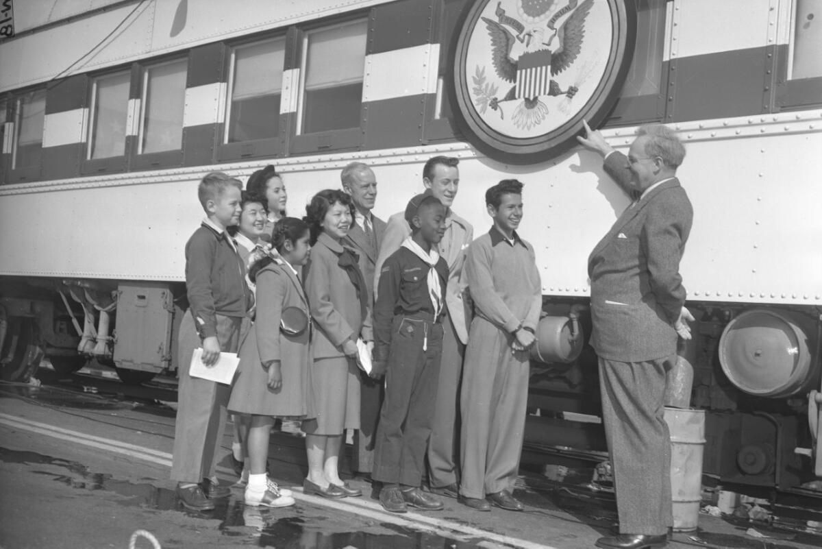 A group of young people and several older men stand next to a train with the U.S. seal on its side.