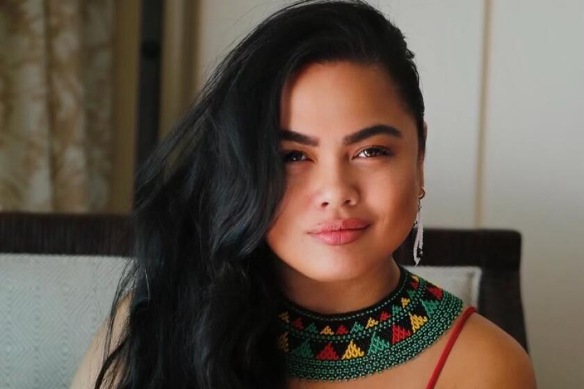 Roslynn Alba Cobarrubias smiles with her hands folded while wearing a red tank top and a colorful patterned neck piece