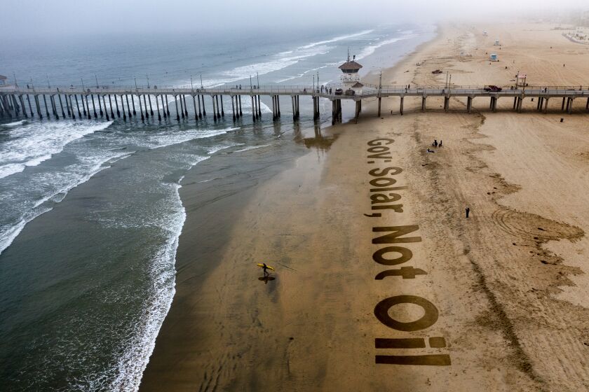Huntington Beach, CA - November 03: A surfer heads out as climate-change and solar activists finish writing a giant 15-foot message in sand, reading "Gov: Solar, Not Oil," at the Huntington Beach pier calling on Gov. Gavin Newsom to increase renewable energies in the wake of last months oil spill in Huntington Beach. Photo taken at the pier at on Wednesday, Nov. 3, 2021 in Huntington Beach, CA. The event was put on by activists from Save California Solar. Organizers are collecting petition signatures ahead of an upcoming proposed decision on the "Net Energy Metering" policy. (Allen J. Schaben / Los Angeles Times)
