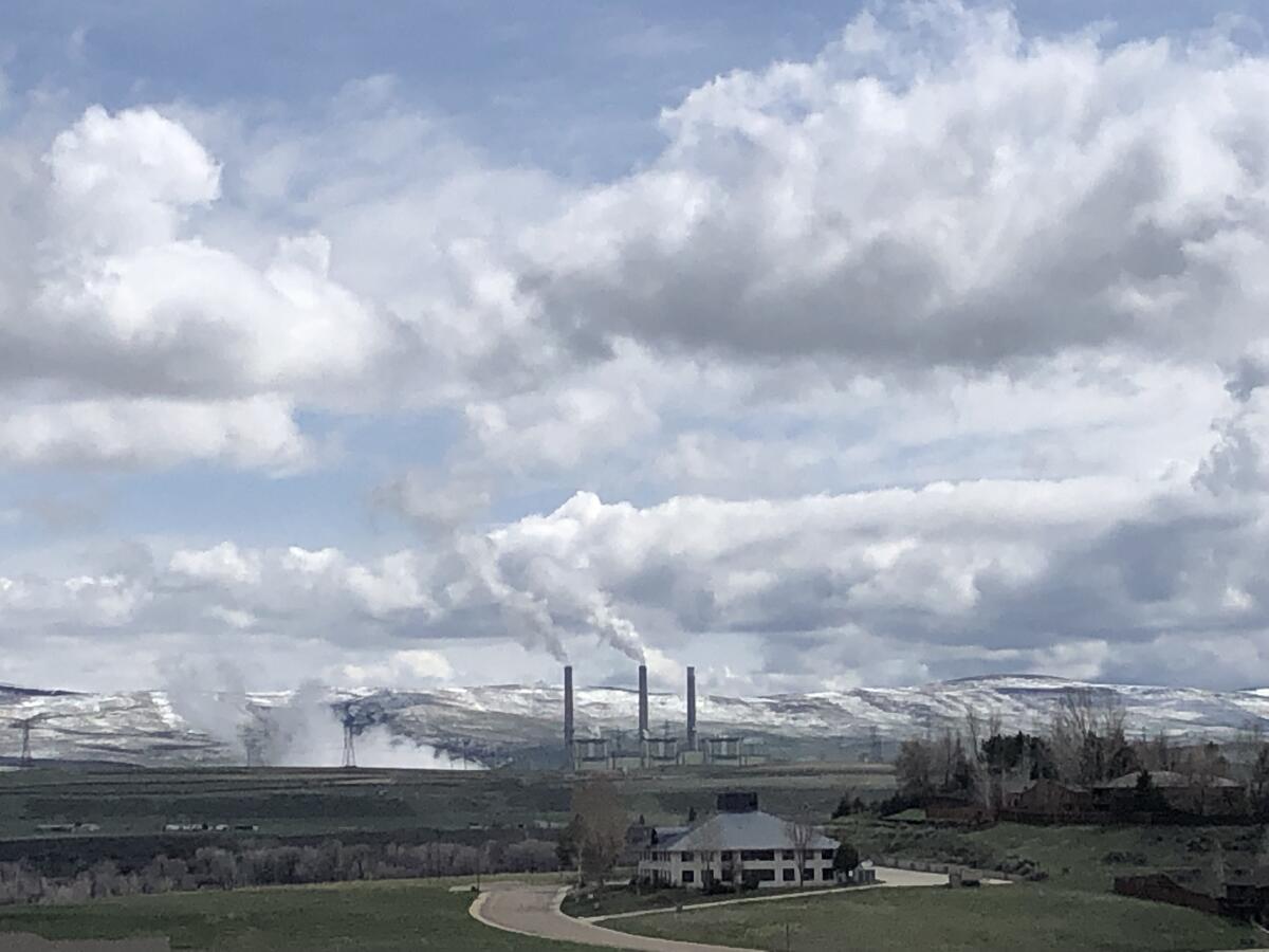 A landscape photo with a multistory building amid fields, three smokestacks belching smoke and low mountains with snow.