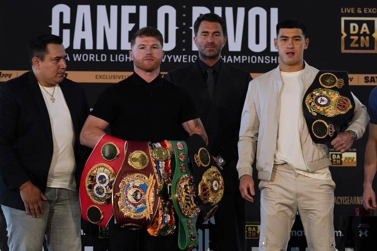 Boxers Canelo Álvarez and Dmitry Bivol pose with their title belts as promoter Eddie Hearn and trainer Eddy Reynoso look on. 