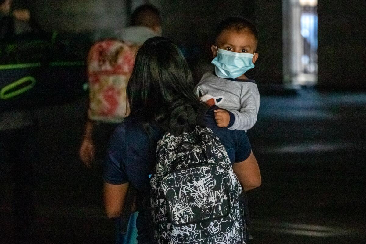 A baby in a mask looks over the shoulder of a woman wearing a backpack.