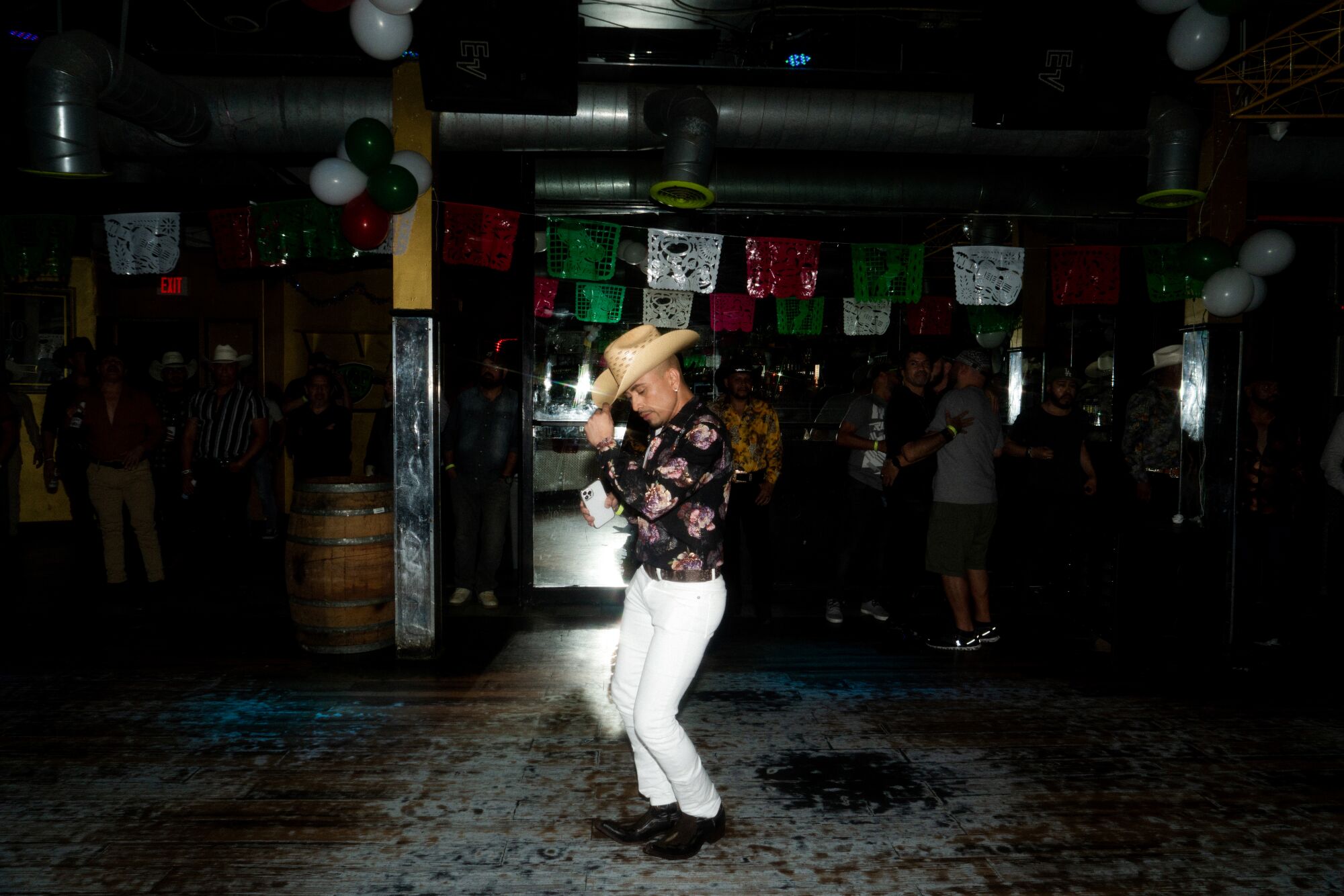 A man in white pants, a flowered shirt and a cowboy hat dances alone on a club dance floor.